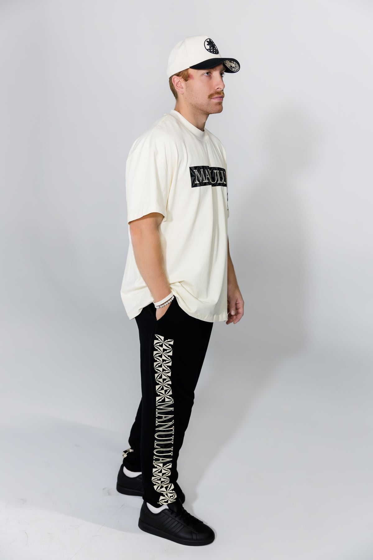 Man wearing a cream hoodie, black pants with white patterns, and a white cap on a gray background