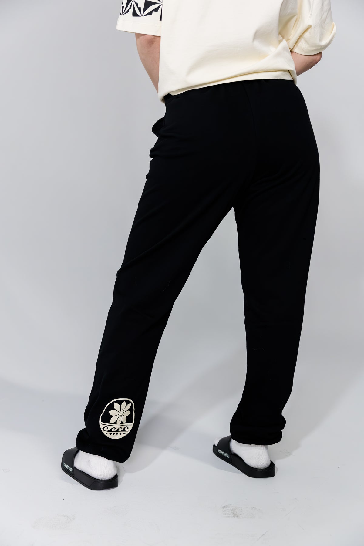 Person wearing black pants with a logo and white shirt on a plain background