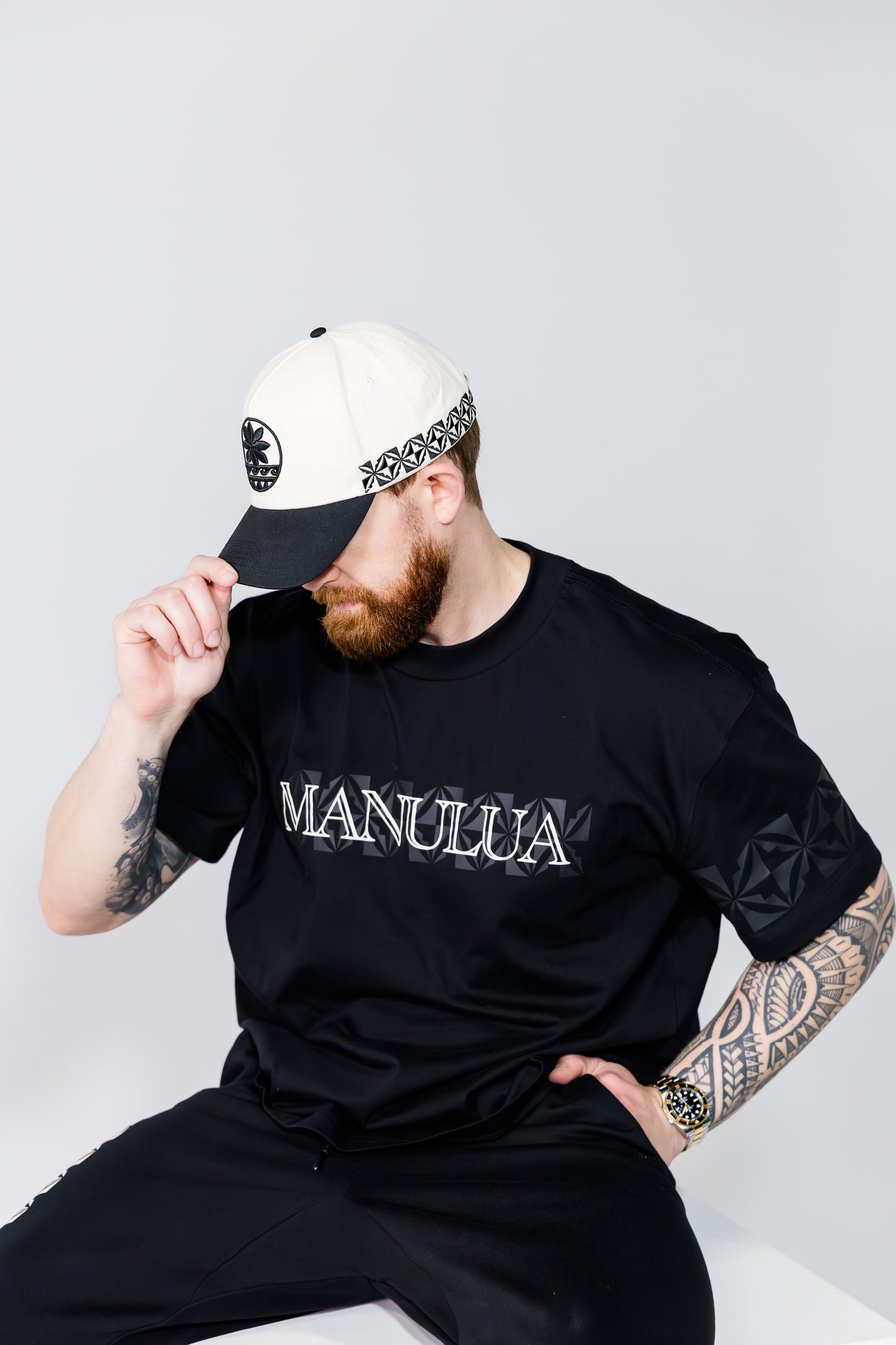Man wearing a black 'MANULUA' t-shirt and white cap with a black and white design.