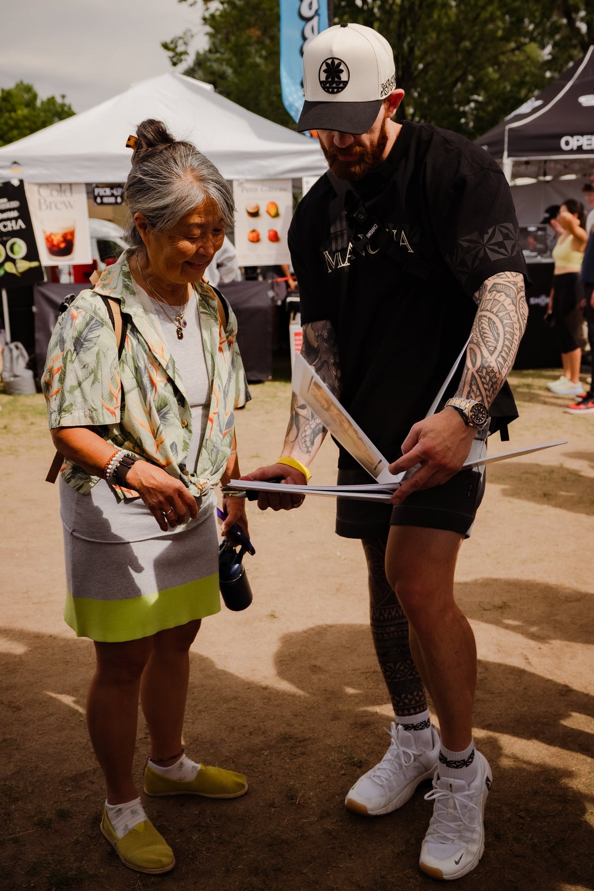 A man and a woman standing outdoors at what appears to be a fair or market, holding papers. The man is wearing a white cap, black t-shirt, black shorts, and white sneakers, while the woman wears a patterned top, a gray skirt, and yellow shoes.