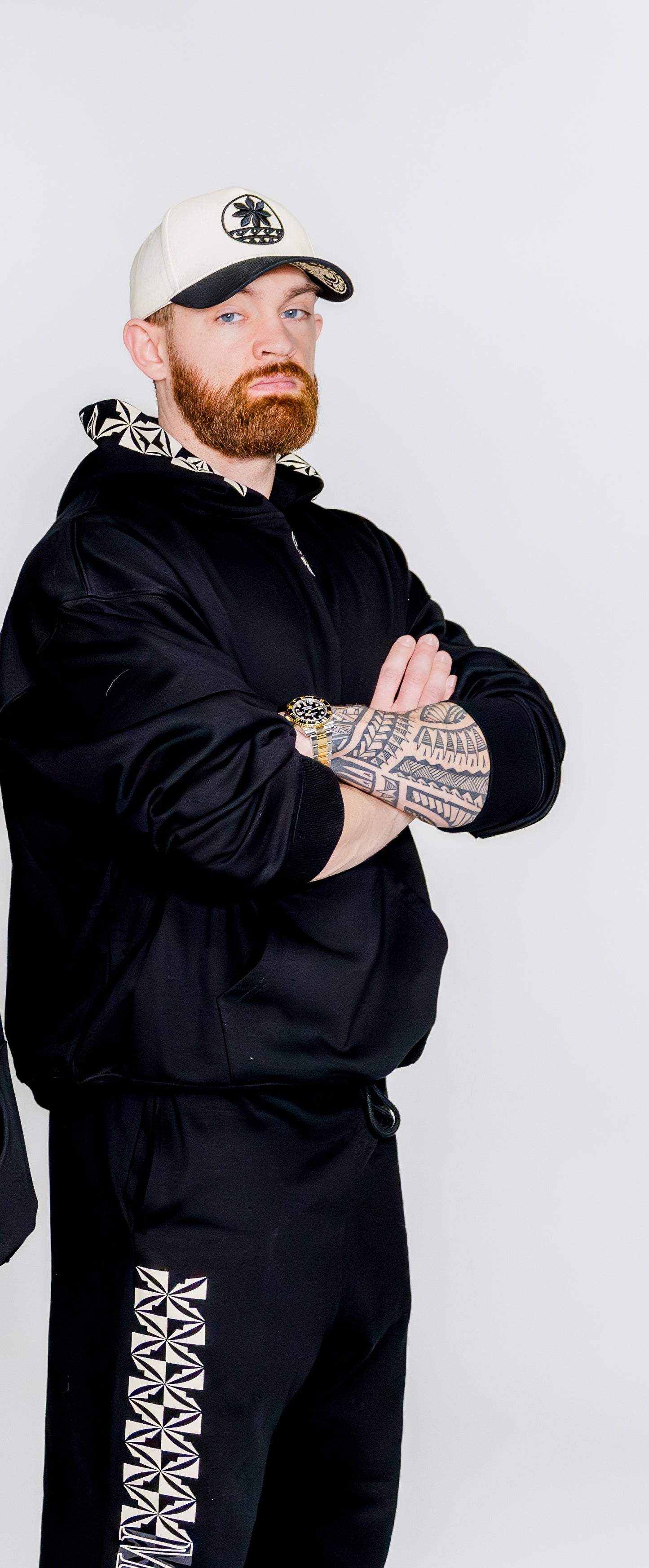 Man wearing a black tracksuit with white patterns and a cap on a light gray background