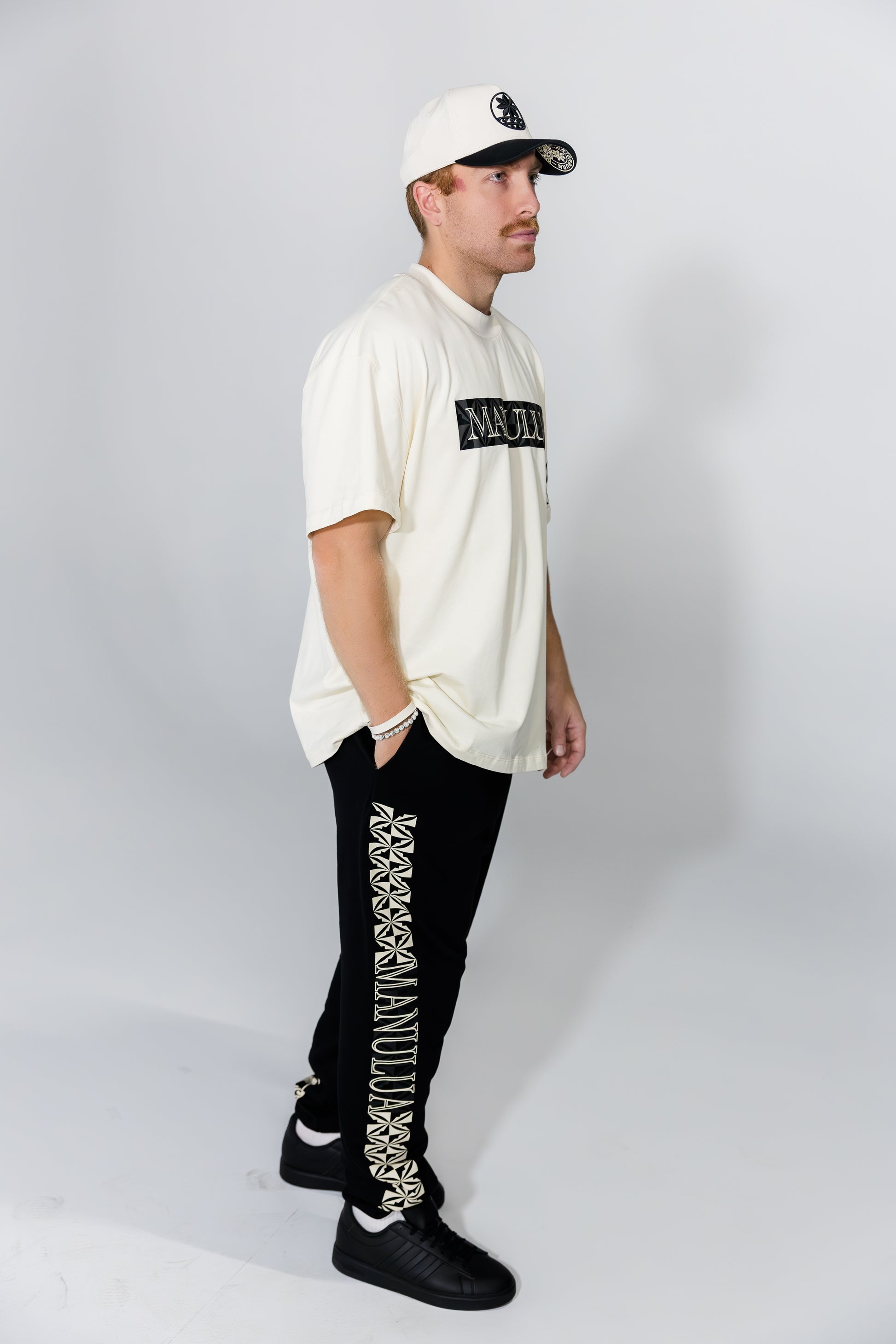 Man wearing a cream hoodie, black pants with white patterns, and a white cap on a gray background