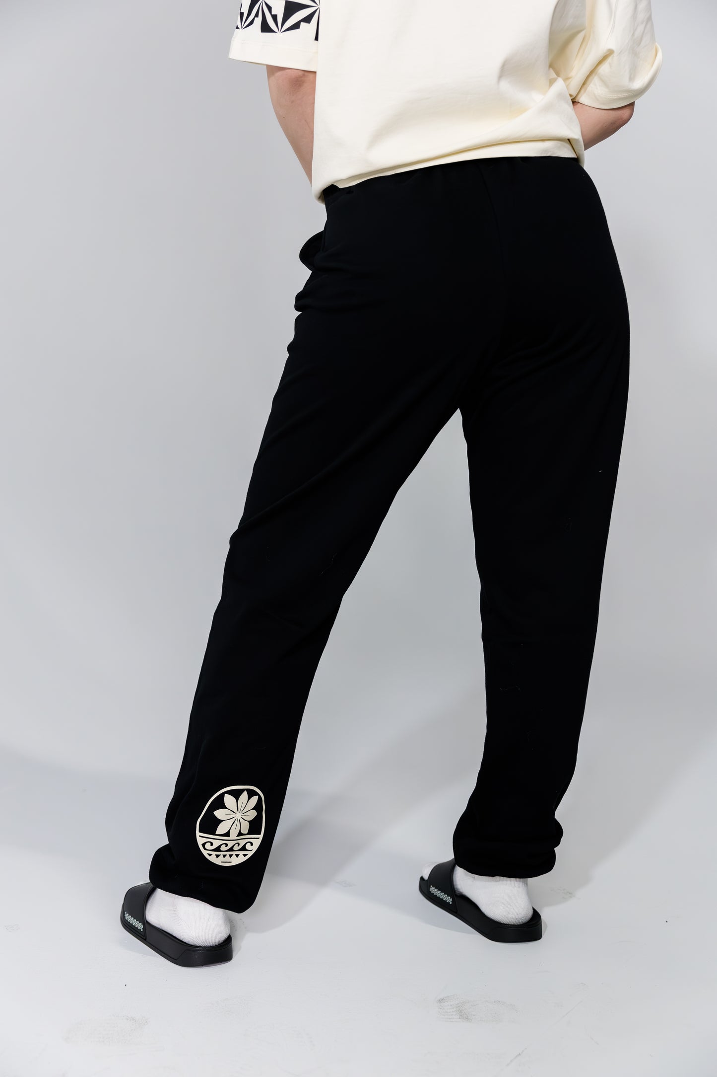 Person wearing black pants with a logo and white shirt on a plain background