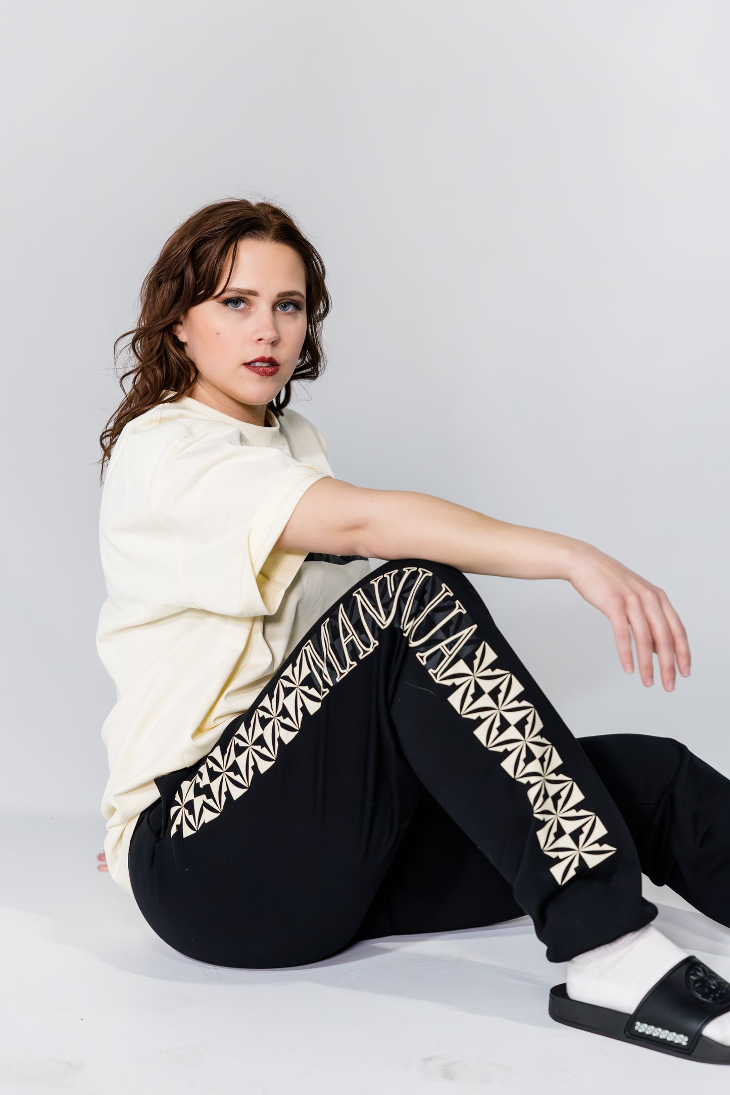 Woman wearing a white top and black pants with geometric patterns, sitting on a light gray background.