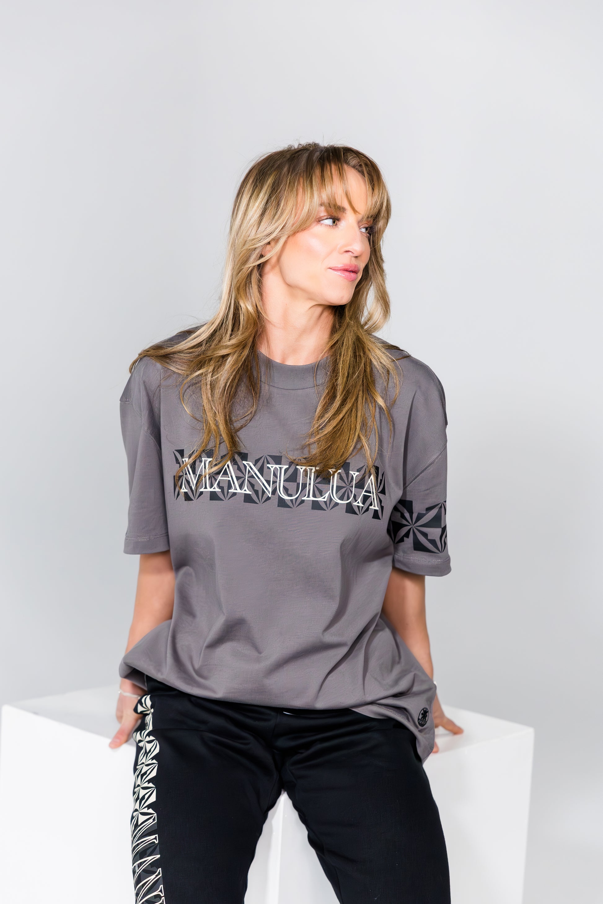 Woman wearing a gray t-shirt with text and graphics, sitting on a white surface against a light gray background.