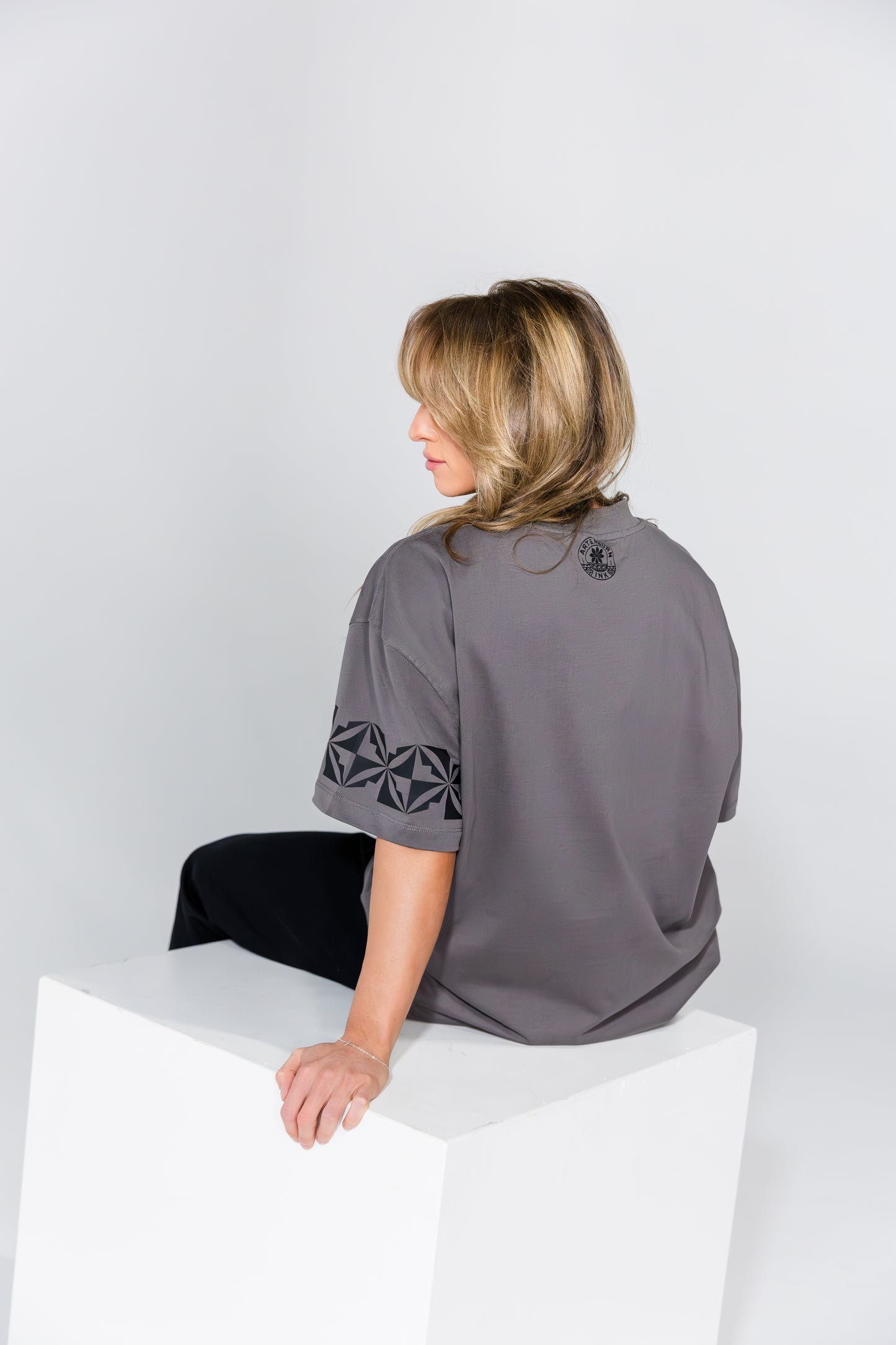 Woman wearing a gray shirt with geometric patterns sitting on a white surface against a light gray background