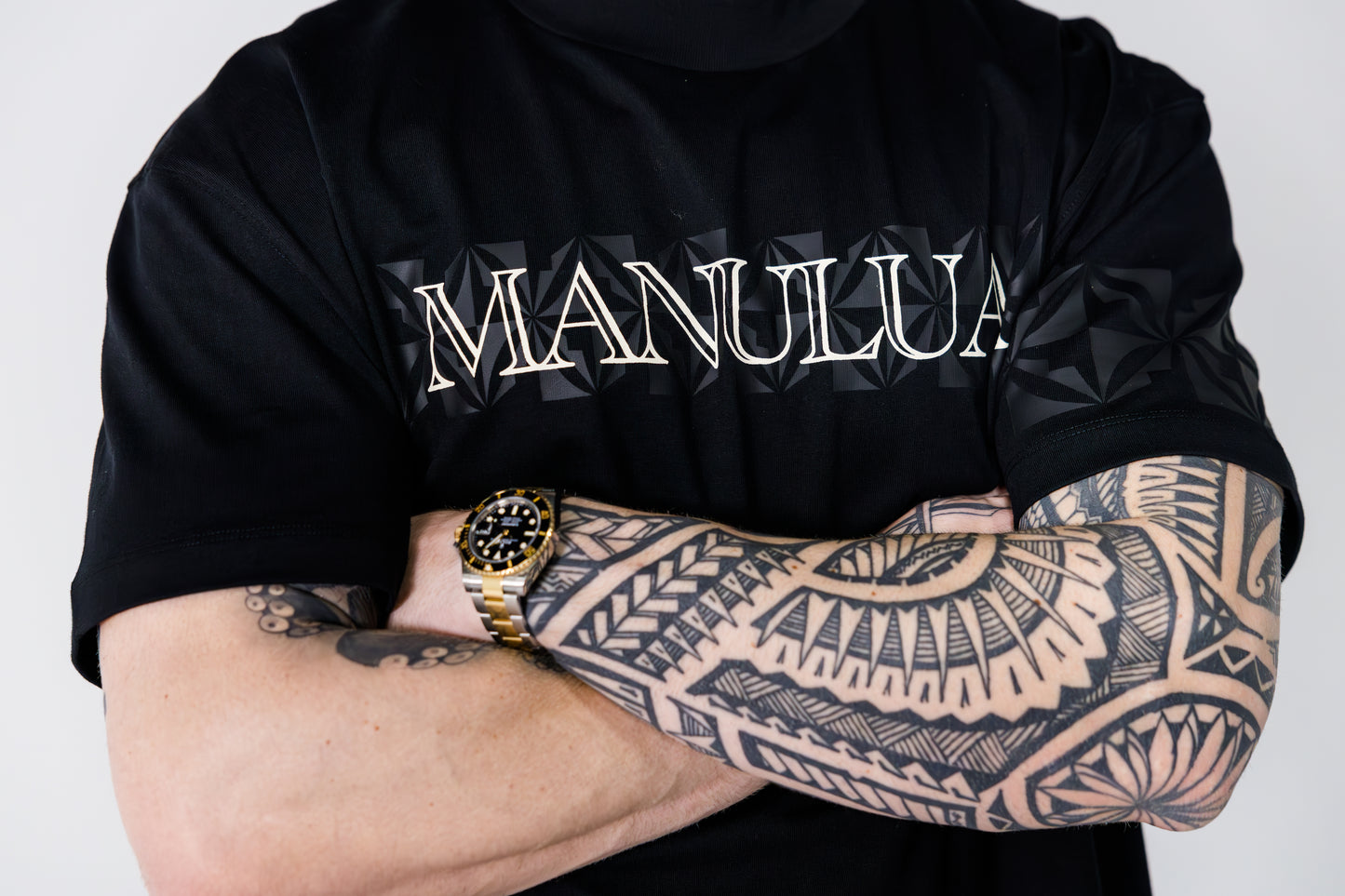 Person wearing a black t-shirt with 'MANULUA' text, displaying tattooed arms and a gold watch.